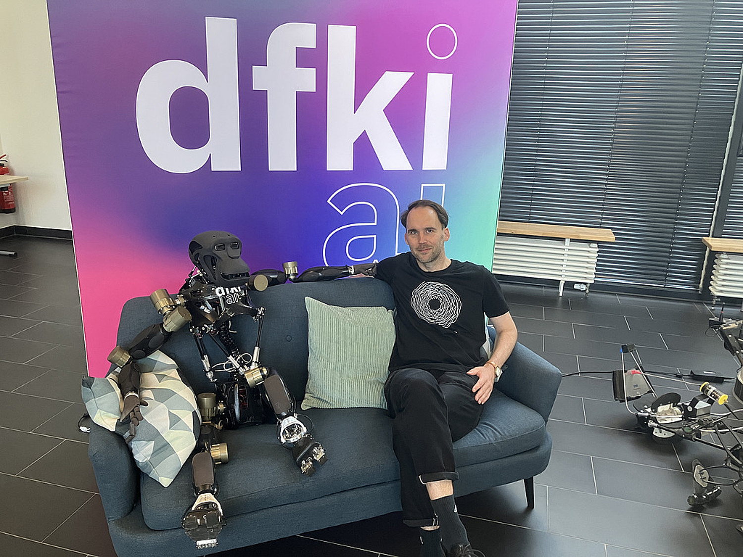 Gereon Rahnfeld sitzt auf einem Sofa neben einem humanoiden Roboter im Robotics Innovation Center des DFKI.